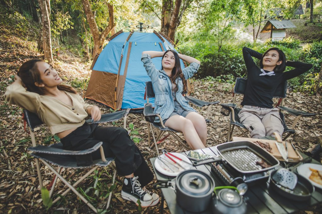 Group of pretty women sitting on camping chair at front of tent for relaxtion while has camping in nature forest with happiness together, copy space