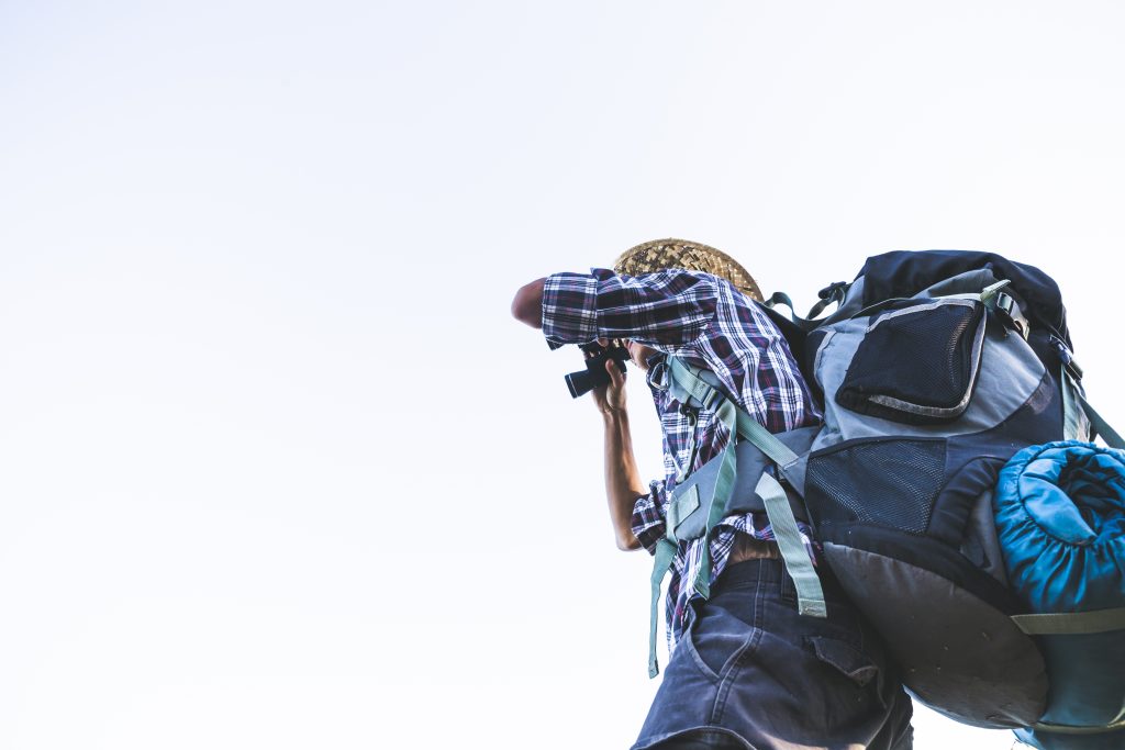 Tourist is watching through binoculars on sunny cloudy sky from mountain top. sun rays. man wear big backpack against sun light