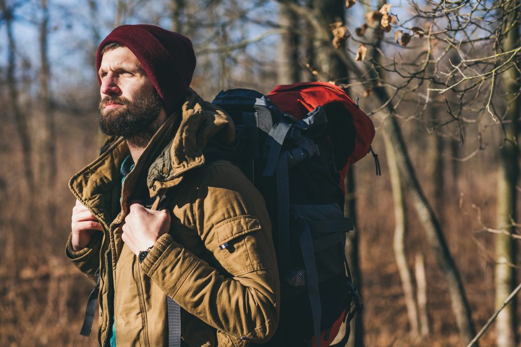 young hipster man traveling with backpack in autumn forest wearing warm jacket and hat, active tourist, exploring nature in cold season