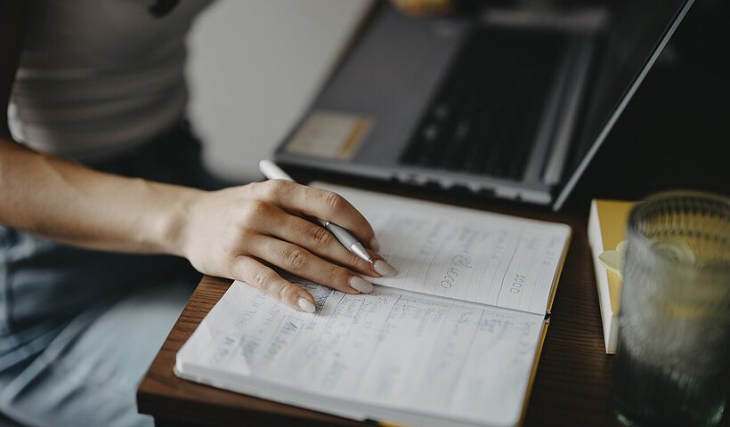 Person writing notes in a notebook beside a laptop at a bright desk