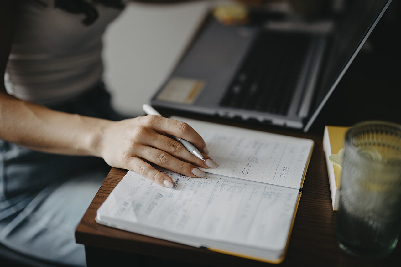 Person writing notes in a notebook beside a laptop at a bright desk